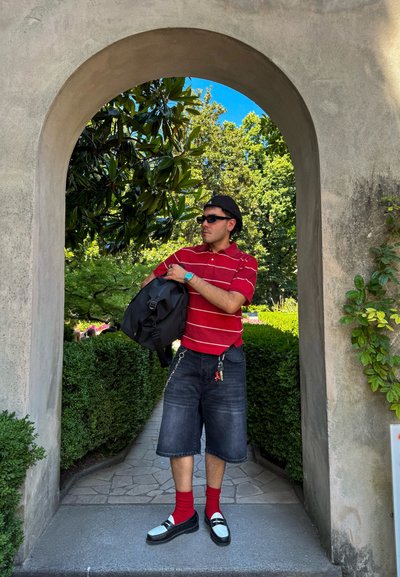 Mochila negra sostenida por una persona que lleva una camiseta de rayas rojas, pantalones cortos de mezclilla, calcetines rojos y zapatos en blanco y negro, enmarcada por un arco.