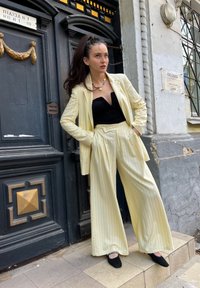 Κρέμα κοστούμι με ρίγες και φαρδιά παντελόνια, συνδυασμένο με εφαρμοστό μαύρο τοπ. Μαύρα μπαλαρίνες και μια κολιέ με χάντρες ολοκληρώνουν την εμφάνιση. Βρίσκεται κοντά σε ένα ιστορικό κτίριο.