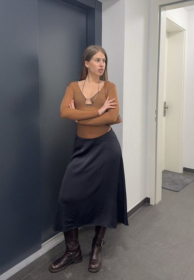 Mujer con blusa marrón, falda negra y botas, de pie con los brazos cruzados junto a una puerta de ascensor oscura cerrada en un pasillo moderno.