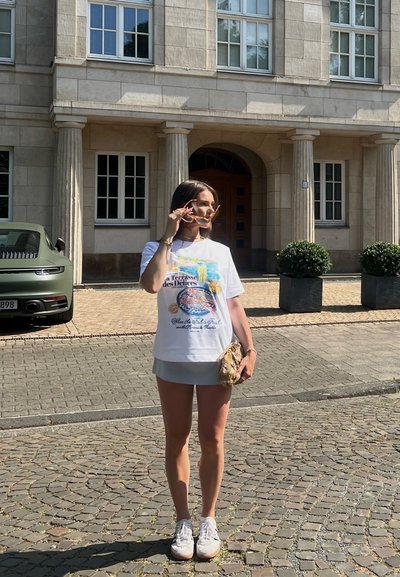 Camiseta gráfica blanca con diseño colorido, falda mini gris y zapatillas blancas. Sosteniendo gafas de sol y un bolso estampado, en un entorno urbano.