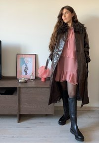 Hnedý kožený trenčkot s kožušinovým golierom cez ružové vrstvené šaty, doplnený čiernymi čižmami po kolená. Moderný dizajn a elegantné spracovanie.