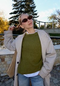 Béžový trenčkot cez zelený pletený sveter, nosený s svetlomodrými džínsami. Hnedé vzorované slnečné okuliare a hladký účes. Vonkajšie prostredie.