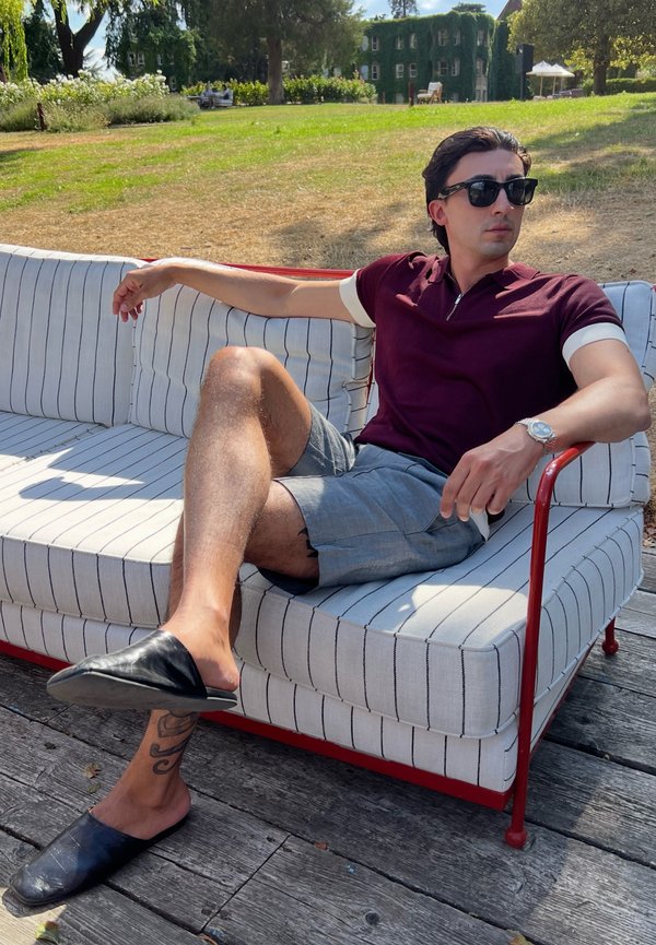 Homme portant des lunettes de soleil, une chemise bordeaux et un short gris assis sur un canapé d'extérieur rayé avec un cadre rouge sur une terrasse en bois dans un parc ensoleillé.