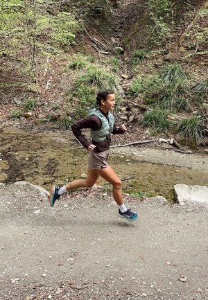 Corridore in un gilet verde chiaro e maglietta a maniche lunghe marrone, con pantaloni corti grigi e scarpe da corsa blu, su un sentiero di terra accanto a un torrente.