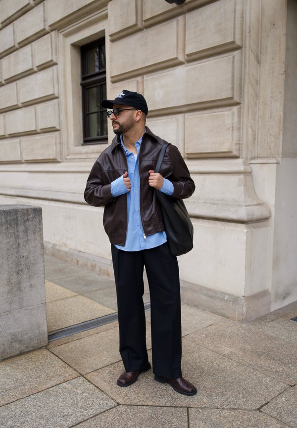 Mann mit schwarzer Kappe, Brille, brauner Lederjacke, blauem Hemd, schwarzen Hosen und Stiefeln, der eine schwarze Tragetasche auf dem Bürgersteig in der Stadt neben einem Gebäude hält.
