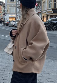 Beige oversized jakke med store knapper og sidelommer, båret med en mørk strikket beanie, kombineret med en cremefarvet håndtaske, på en gade i byen.