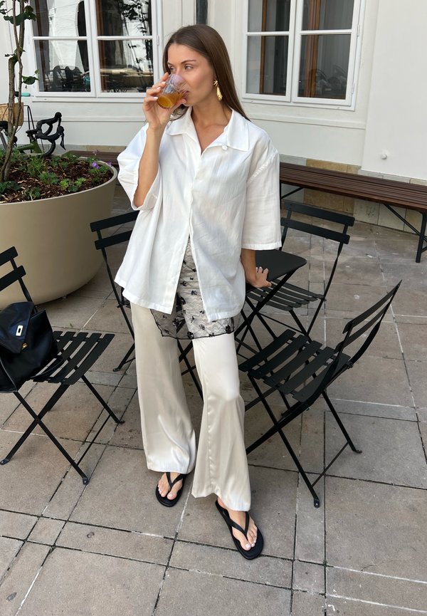 Kvinde i hvid bluse og bukser drikker af et glas, mens hun står nær sorte metalstole og en stor plantekasse på en flisebelagt terrasse.