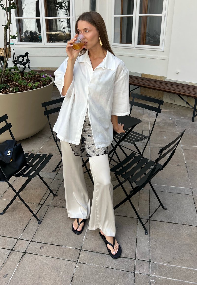 Woman in white blouse and pants drinks from a glass while standing near black metal chairs and a large planter on a tiled patio.