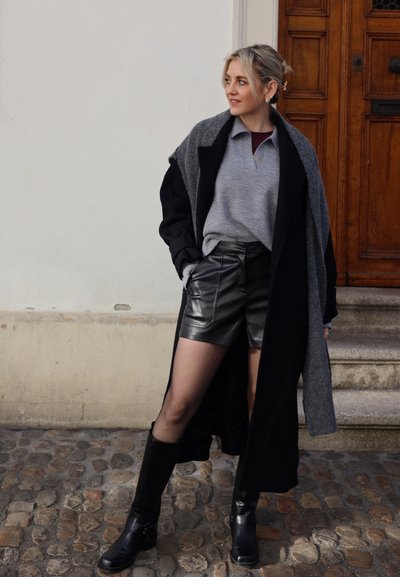 Abrigo largo negro sobre un suéter gris de punto, combinado con shorts de cuero negro de talle alto y botas negras hasta la rodilla. Fondo de camino de piedra.