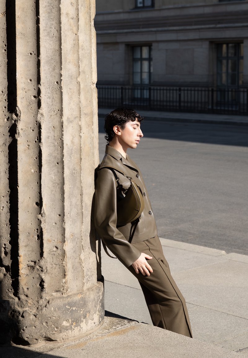 Personne en costume vert olive s’appuie contre une grande colonne en pierre texturée, debout à l’extérieur près d’un bâtiment avec des fenêtres grillagées en plein jour.