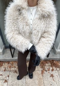 Roomkleurige nepbontjas met een brede kraag, gedragen over een wit hemd met knopen, gecombineerd met bruine wijde broekspijpen en zwarte handschoenen.