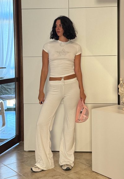 Camiseta blanca de manga corta con un diseño de estrella de strass, combinada con pantalones de campana blancos. Sosteniendo una gorra rosa, de pie contra una pared blanca.