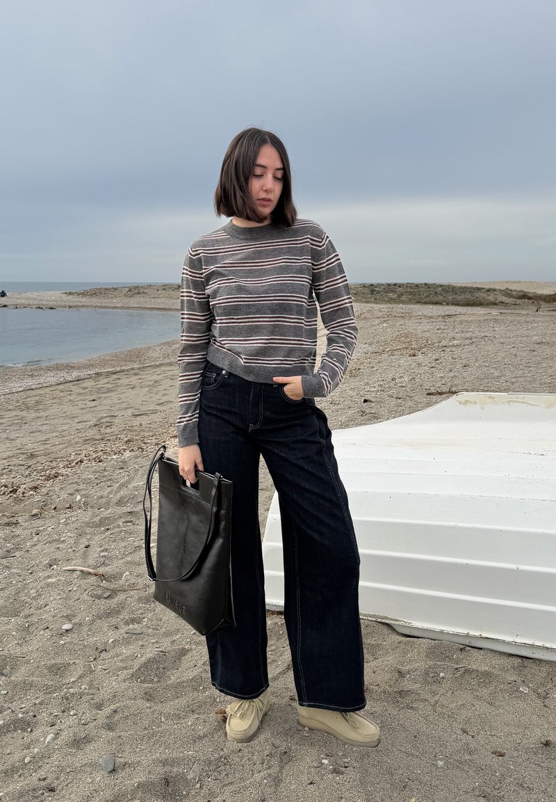 Young woman in striped sweater and wide-leg jeans holds black bag standing on a sandy beach near an upside-down white boat.