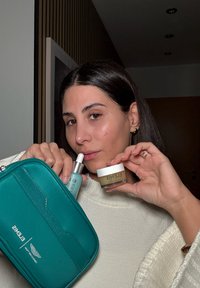 Mujer con cabello oscuro en suéter blanco sosteniendo una bolsa cosmética color teal, una botella de suero azul y un tarro de bálsamo limpiador en interiores con una pared de paneles de madera detrás.