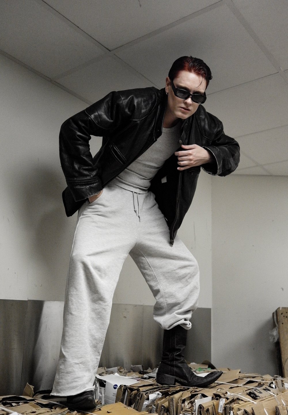 Veste en cuir noire, sweat-shirt gris, pantalon de jogging gris ample et bottes noires. Pose sur des cartons dans un environnement intérieur.