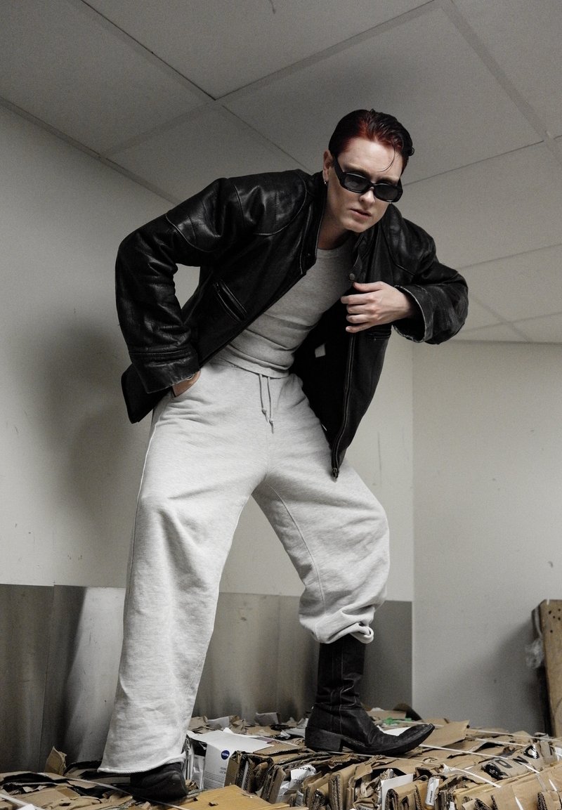 Veste en cuir noire, sweat-shirt gris, pantalon de jogging gris ample et bottes noires. Pose sur des cartons dans un environnement intérieur.