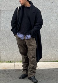 Μαύρο μάλλινο παλτό, υφασμάτινος μαύρος σκούφος, επικαλυμμένος με καρό μπλε πουκάμισο, ελαιόχρωμα cargo παντελόνια και μαύρες μπότες. Στηρίζεται σε έναν γκρίζο τοίχο.