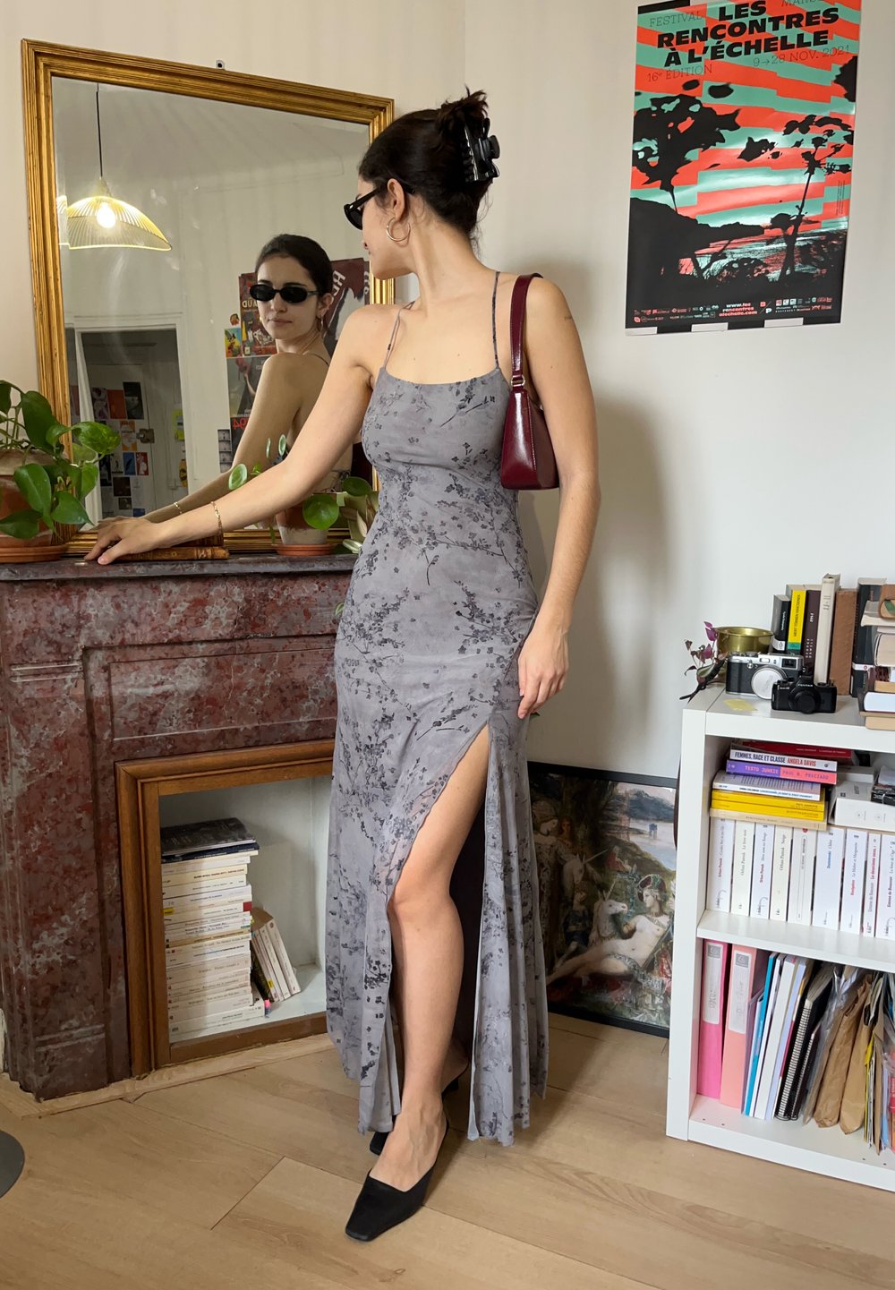 Femme en robe grise à motifs floraux avec une fente sur la cuisse et des chaussures noires regardant son reflet dans un miroir placé au-dessus d'une cheminée en marbre rouge.