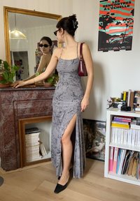 Femme en robe grise à motifs floraux avec une fente sur la cuisse et des chaussures noires regardant son reflet dans un miroir placé au-dessus d'une cheminée en marbre rouge.