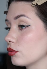 Visage d'une femme de profil avec un eye-liner ailé, un rouge à lèvres rouge, un anneau au nez, et les cheveux retenus en arrière par des pinces beige.