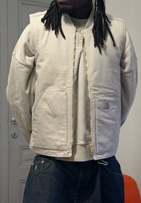 Beige väritetty kangastakki, jossa on vetoketjullinen suljin, kaksi etutaskua ja pieni logoplässi. Takissa on resorikaulus ja ompelu-yksityiskohdat.