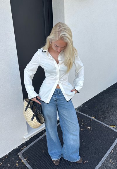 Camisa blanca de manga larga con botones y puños negros con forma de concha, combinada con jeans de pierna ancha azul y un bolso tejido en negro y beige.
