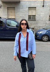 Μπλε πουκαμίσα, μπορντώ γραβάτα, σκούρο τζιν και τσαντάκι με σχέδιο. Γυαλιά ηλίου και μονόχρωμο φόντο με παρκαρισμένα αυτοκίνητα και πέτρινα κτίρια.