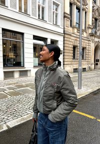 Graue Bomberjacke mit strukturierten Vorderdetails und hohem Kragen, kombiniert mit blauen Jeans. Schwarze Handtasche sichtbar. Urbane Umgebung.