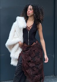 Musta hihaton toppi vetoketjulla, kerroksittain oleva ruskea pitsihame kukkakuviolla ja valkoinen tekoturkistakki. Asua koristaa kultainen kaulakoru.