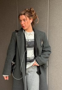 Mladá žena s slúchadlami, oblečená v sivej kabáte, pruhovanej košeli a sivých nohaviciach, stojaca pri textúrovanom múre, pozerajúca sa doľava.