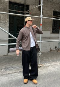 Brūna un balta svītraina pogām aiztaisāma krekls, balta T-krekls zem tā, melnas plašas bikses un brūnas zamša kurpes, rokās tur dzērienu pelēkā vidē.