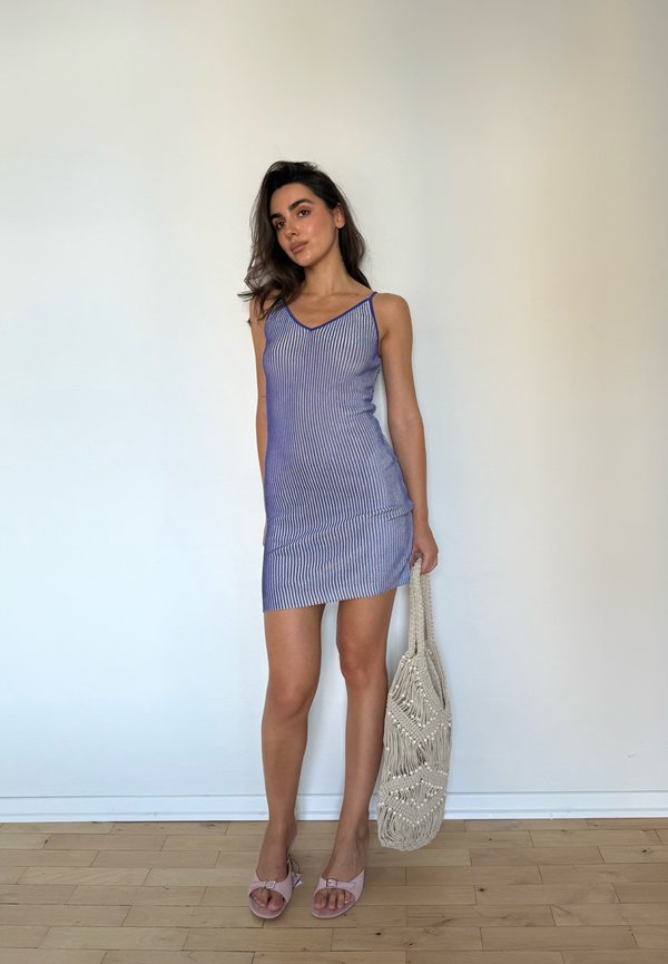 Vrouw staande in een blauw gestreepte mouwloze jurk, houdend een gevlochten beige tas, met roze open-toe sandalen op een houten vloer.
