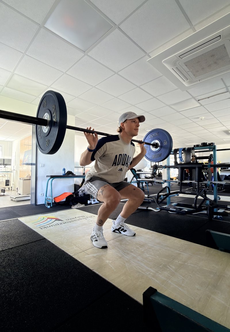 Jeune homme en tenue de sport en train de faire un squat avec une barre chargée sur les épaules dans une salle de sport lumineuse équipée pour l'haltérophilie.