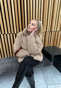 Beige, plysch, oversized jacka med hög krage och brun accentband, tillsammans med svarta knälånga stövlar i krokodilsmönster.