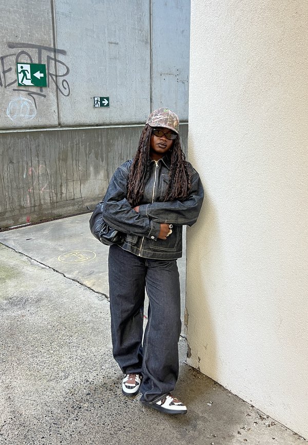 Jeansjacke und Schlaghose in dunkler Waschung, kombiniert mit einer Camouflage-Kappe und schwarz-weißen Turnschuhen. Stehend vor einer Wand.