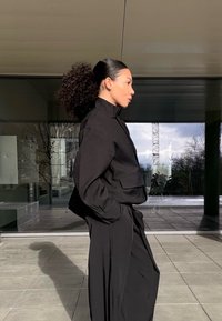 Sort jakke med krave og lange ærmer samt lappelommer, kombineret med løstsiddende sorte bukser, båret af en person i profil foran en glasbaggrund.