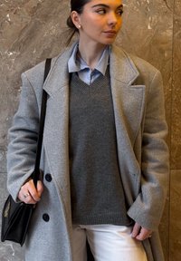 Jeune femme portant un manteau gris, un pull gris foncé, une chemise bleu clair, un pantalon blanc, et portant un sac à bandoulière noir, debout devant un mur en marbre.