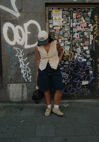 En person i en beige ermeløs vest og mørke denimshorts står mot en vegg dekket av graffiti, holder en mønstret veske og har på seg en caps.