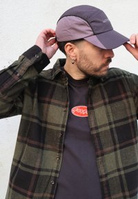 Casquette grise à visière courbée, chemise en flanelle à carreaux verts et noirs portée sur un t-shirt noir avec un logo rouge, avec une apparence de tissu texturé.