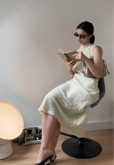 Vestido verde claro, hasta la rodilla, con tela texturizada. Sentada en una silla gris, sosteniendo un libro, usando gafas de sol negras y tacones.