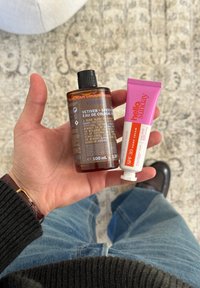 Hånd som holder en brun flaske med Vetiver + Spices eau de cologne og et rosa rør med Hello Sunday SPF 30 håndkrem.