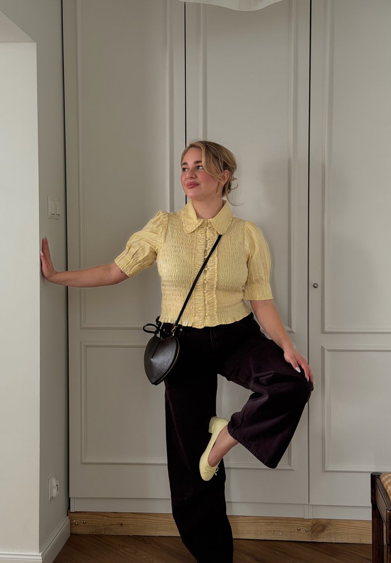 Femme en blouse jaune et pantalon noir, en équilibre sur une jambe avec le bras appuyé contre un mur, portant un sac en forme de cœur et des ballerines jaunes.