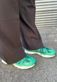 Πράσινα αθλητικά παπούτσια με υφή στο πάνω μέρος, με μοβ λεπτομέρειες και λευκές σόλες. Συνδυασμένα με σκούρους, φαρδιούς παντελόνες.