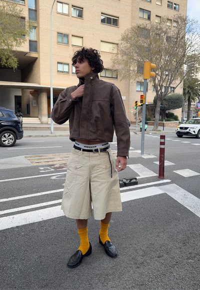 Joven ajustándose la chaqueta marrón mientras está de pie en un paso de peatones de la ciudad, vistiendo pantalones cortos beige, calcetines amarillos y zapatos negros.