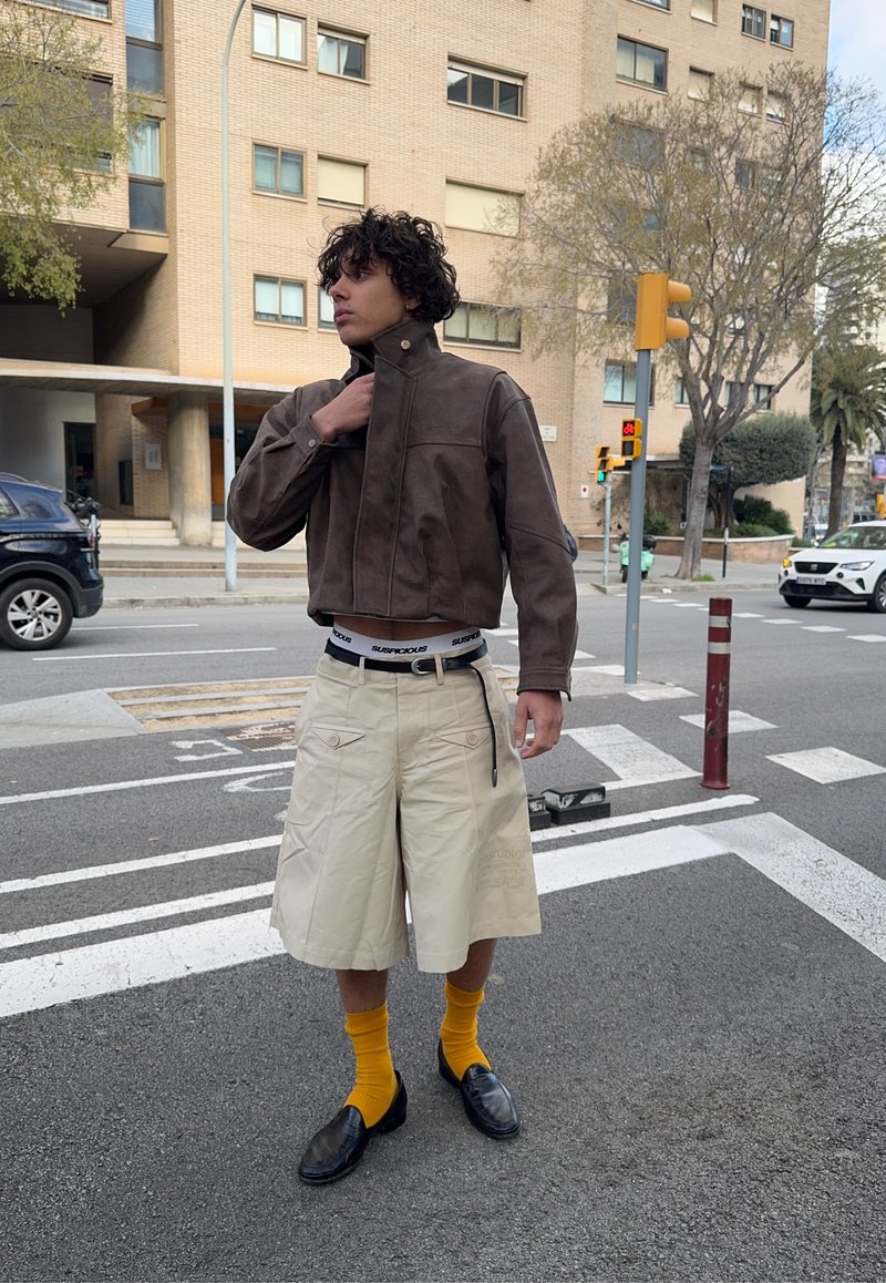 Jeune homme ajustant une veste marron debout sur un passage piéton en ville, portant un short beige, des chaussettes jaunes et des chaussures noires.