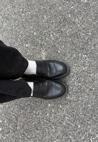 Mocassins de couro preto com uma superfície lisa, apresentando duas faixas horizontais na parte da frente. Usados com meias cinzas num pavimento texturizado.