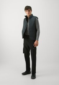 Czarna pikowana kamizelka z wysokim kołnierzem na szarym, długim rękawie. Dobrana z czarnymi spodniami cargo z bocznymi kieszeniami i czarnym obuwiem.