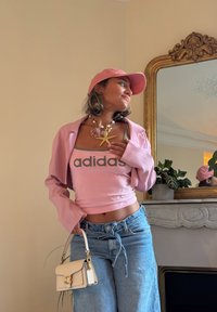 Donna con un top Adidas rosa, giacca e cappellino coordinati, jeans blu, che tiene una borsa bianca, in piedi vicino a uno specchio ornato e a una pianta su un camino.