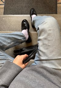 Svart krokodilmönstrad handväska med dragkedja, ljusgrå jeans och polerade svarta loafers i kombination med ljusa strumpor. En avslappnad grå tröja är synlig.