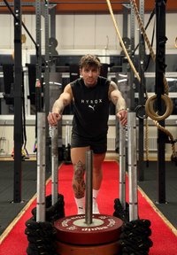 Homme tatoué poussant un traîneau lesté sur une piste rouge dans une salle de sport, entouré de cordes, d'anneaux et d'équipements d'exercice.